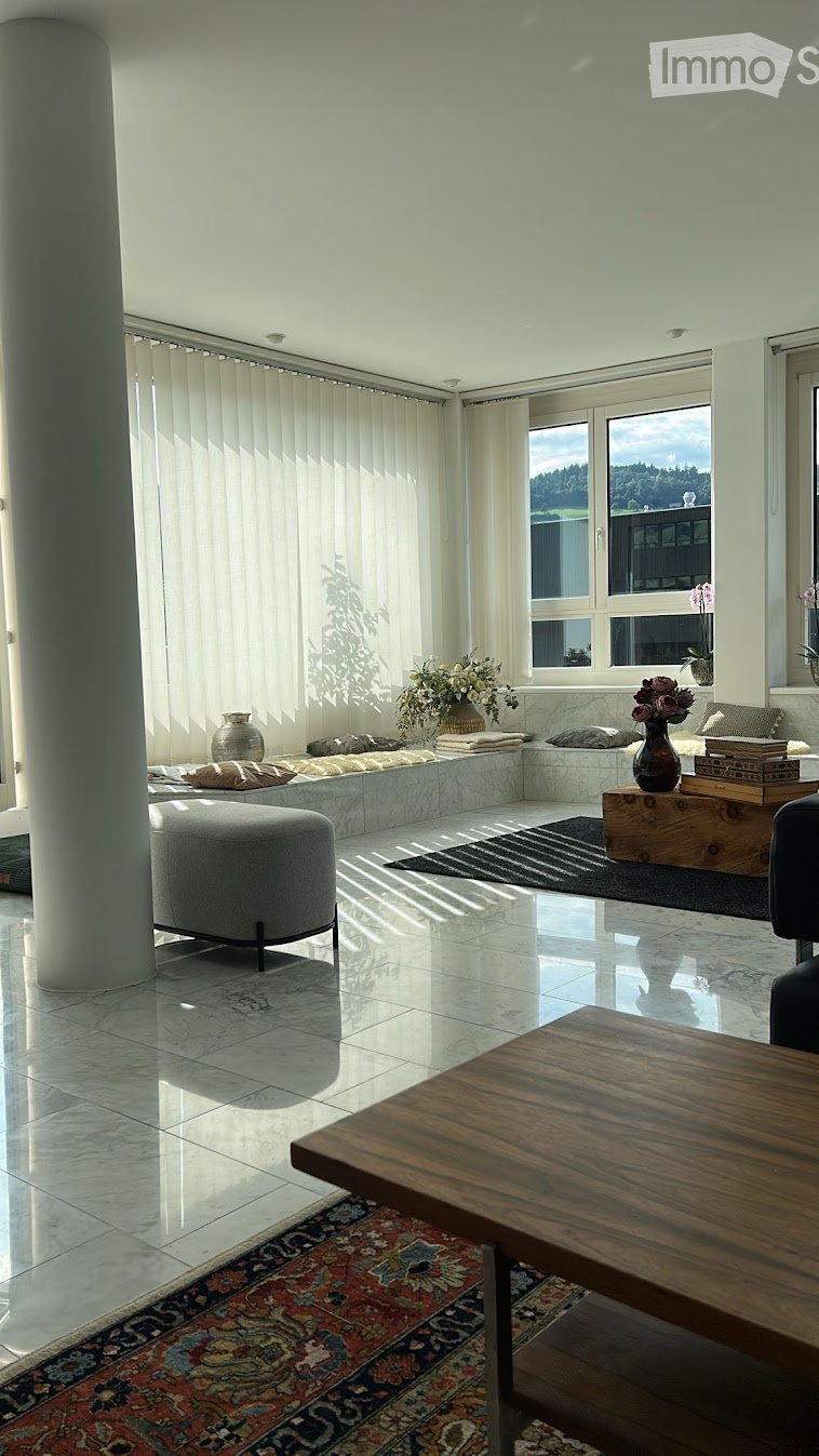 Bright living room with large windows, white walls, polished tile floor, and a mix of modern and traditional furniture including a sofa, coffee table, ottoman, and decorative vases.