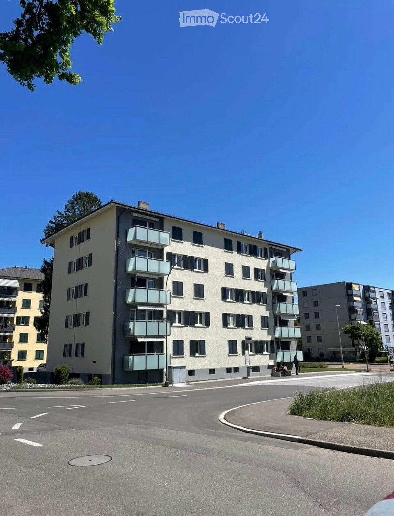 Four-story residential building with multiple balconies and windows, surrounded by other buildings and greenery.