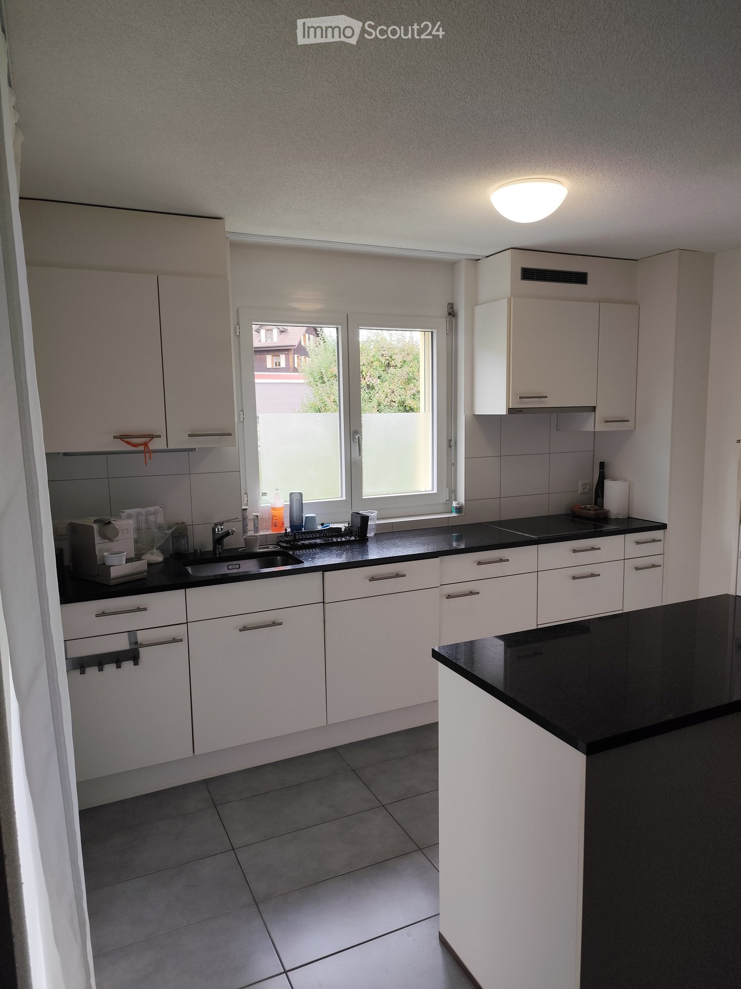 Modern kitchen with white cabinets, black countertops, and a window overlooking a garden. Includes a sink, coffee maker, and various small items on the counter.