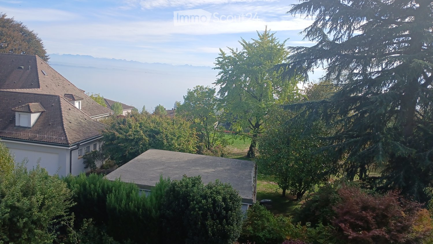 A two-story house with a tiled roof and white exterior, surrounded by lush greenery and trees, with a scenic view of a lake and distant mountains.