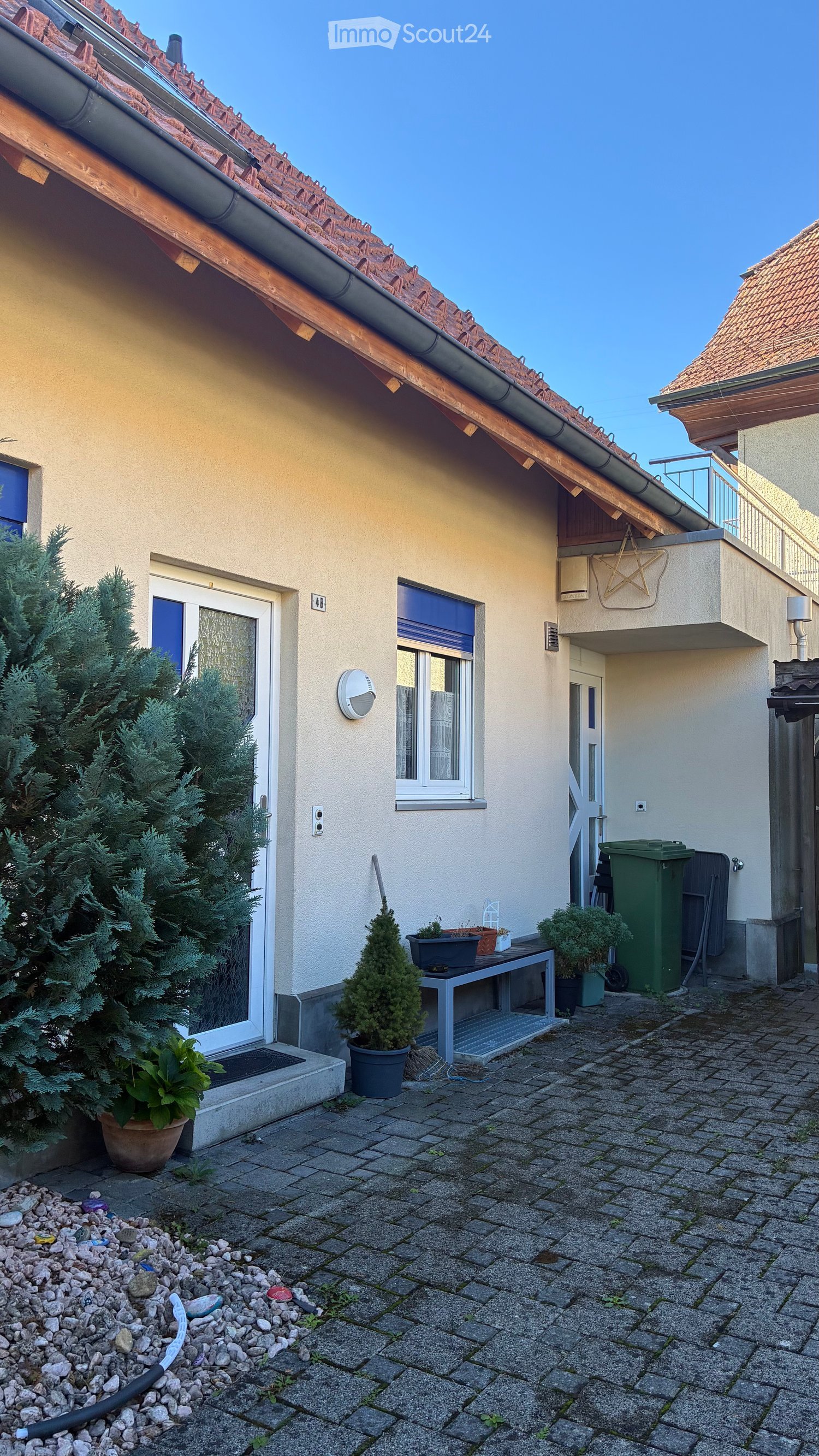 Outdoor patio area with a small table, potted plants, and a trash bin, adjacent to a house with a tiled roof and windows.