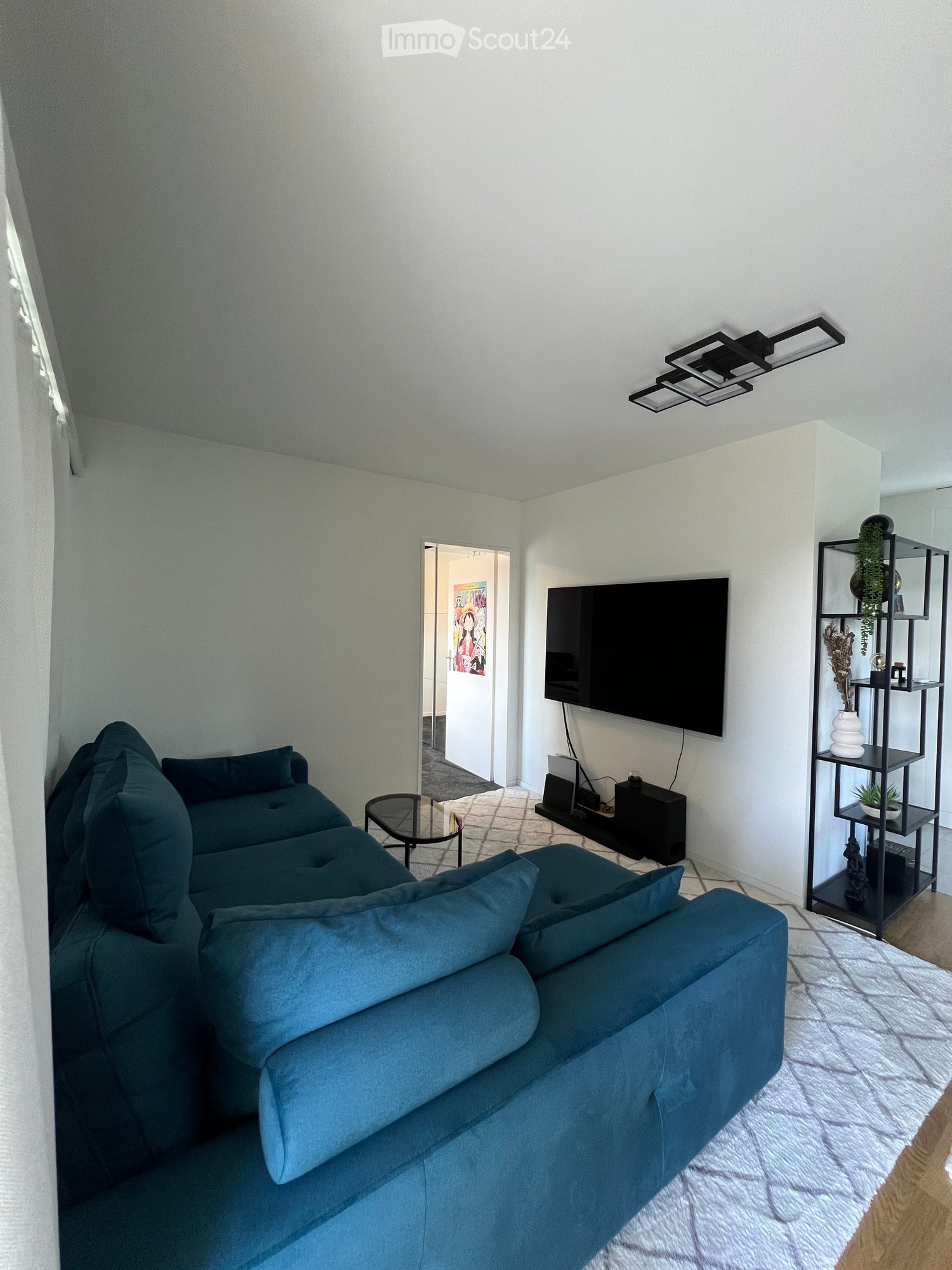 Modern living room with a blue sectional sofa, glass coffee table, flat-screen TV mounted on the wall, and a black shelving unit with decorative items and plants.