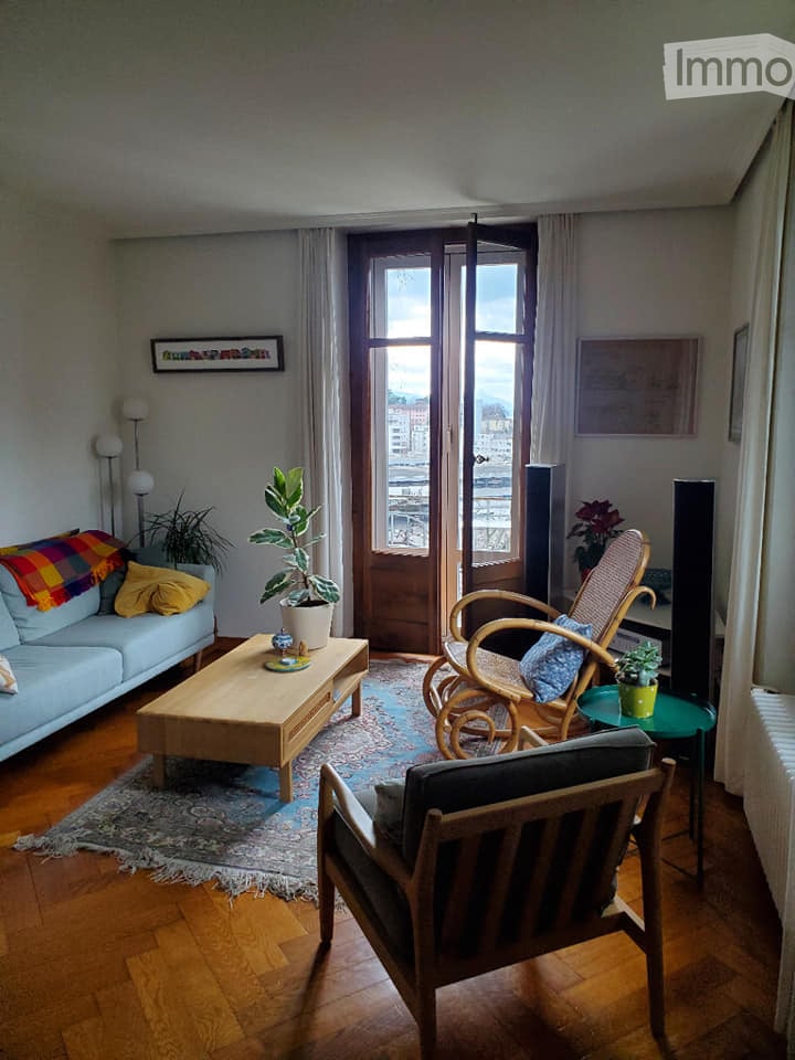 Bright living room with wooden flooring, large windows offering a view, a light blue sofa with colorful cushions, a wooden coffee table, a wicker hanging chair, and a wooden armchair.