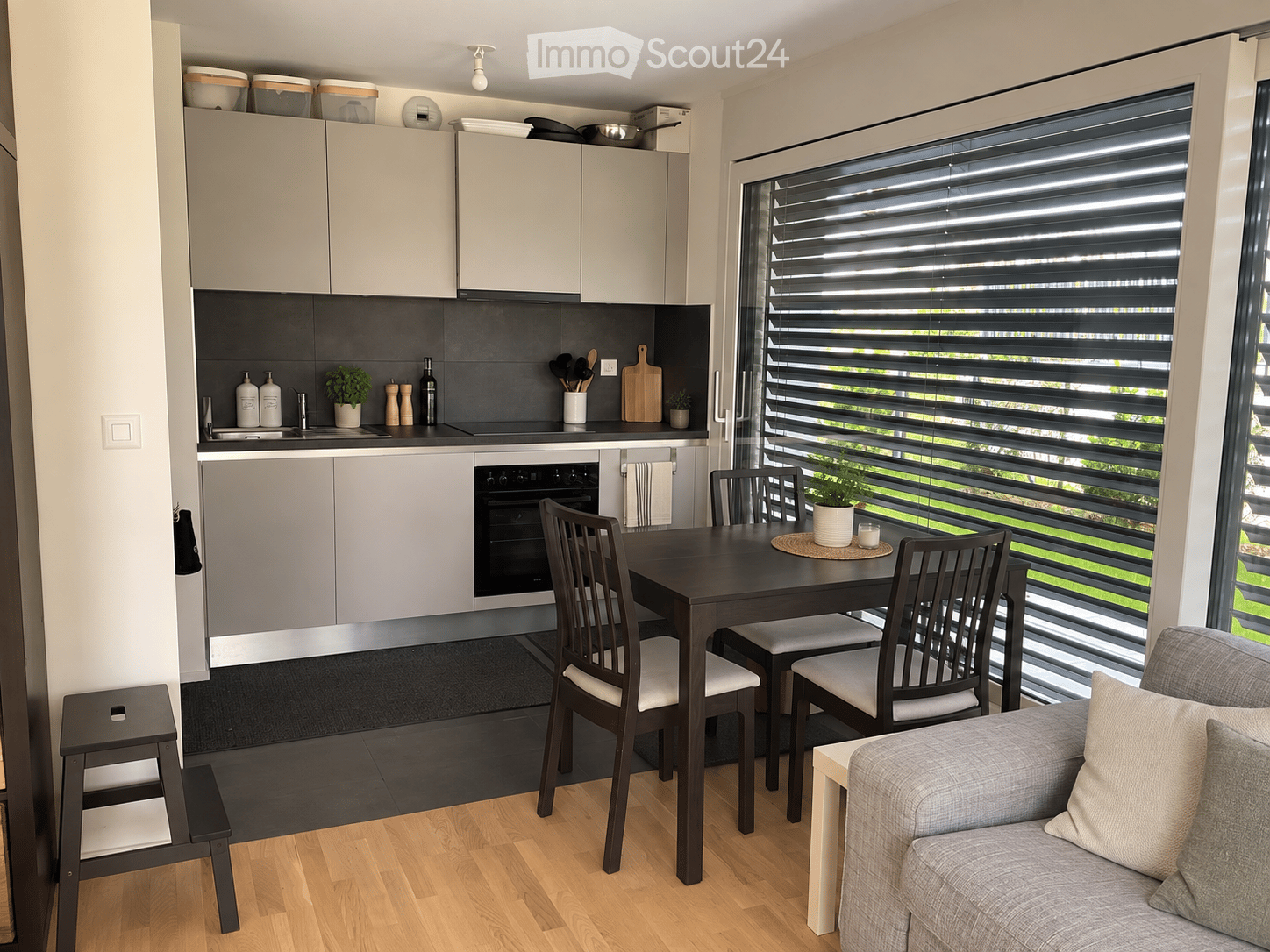 Modern, open-plan living and dining area with grey sofa, dining table with chairs, kitchen cabinets, and appliances.