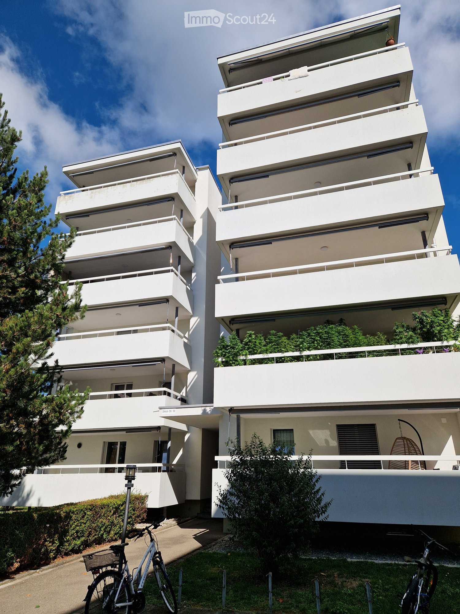 Modern white apartment building with multiple balconies and greenery, bicycles parked in front.