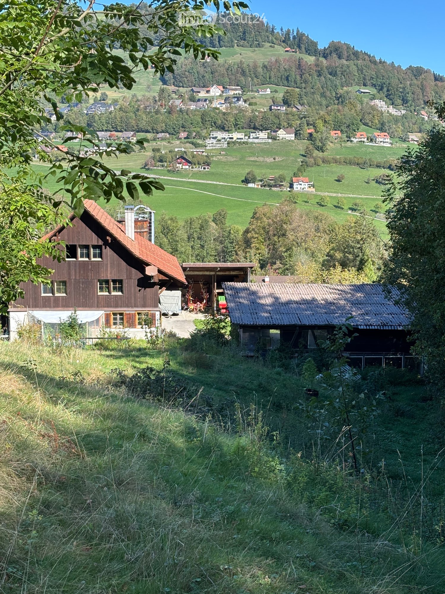 A scenic rural landscape with a wooden house, a small shed, and a view of a valley with scattered houses and green fields.