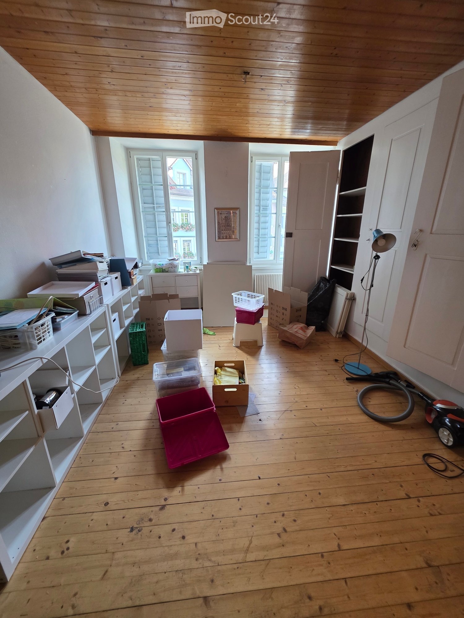 Empty room with wooden flooring, white walls, and large windows. The space contains storage boxes, a vacuum cleaner, and a floor lamp, but no furniture.