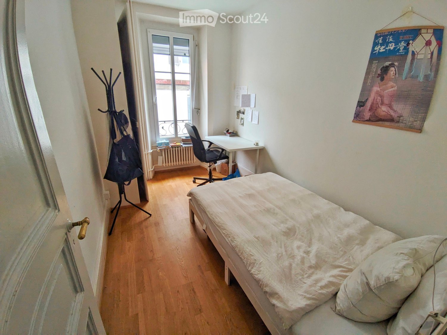 A small, furnished bedroom with a single bed, desk, chair, coat rack, and a poster on the wall. The room has wooden flooring and a window with a view of the street.