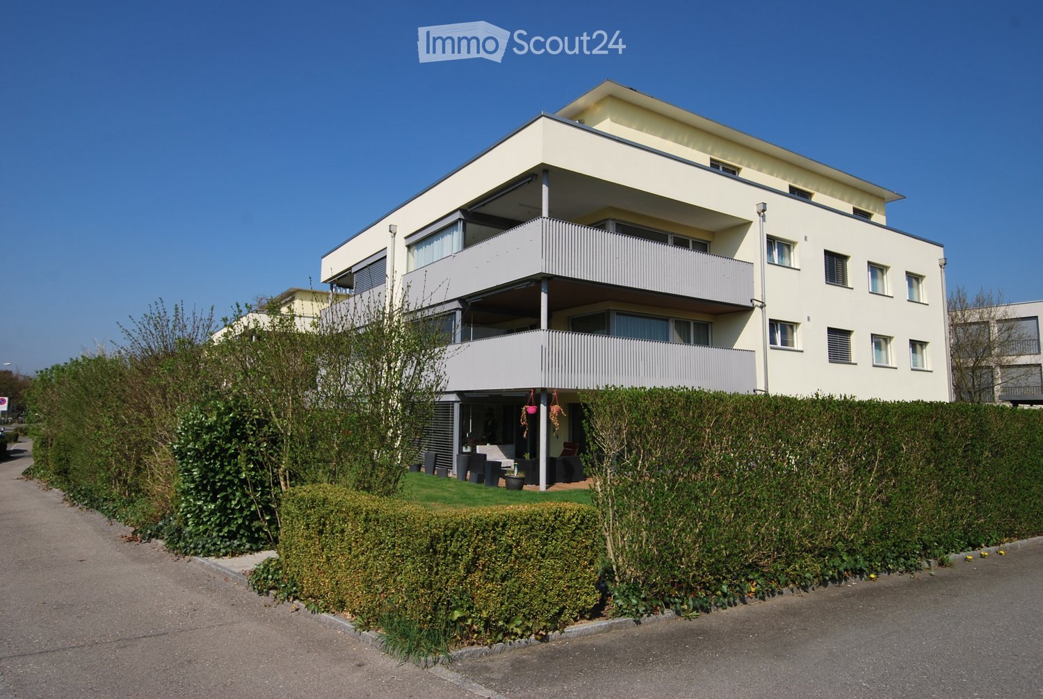 Modern two-story residential building with white exterior, large windows, and balconies. The ground floor features a small patio area with outdoor furniture and plants.