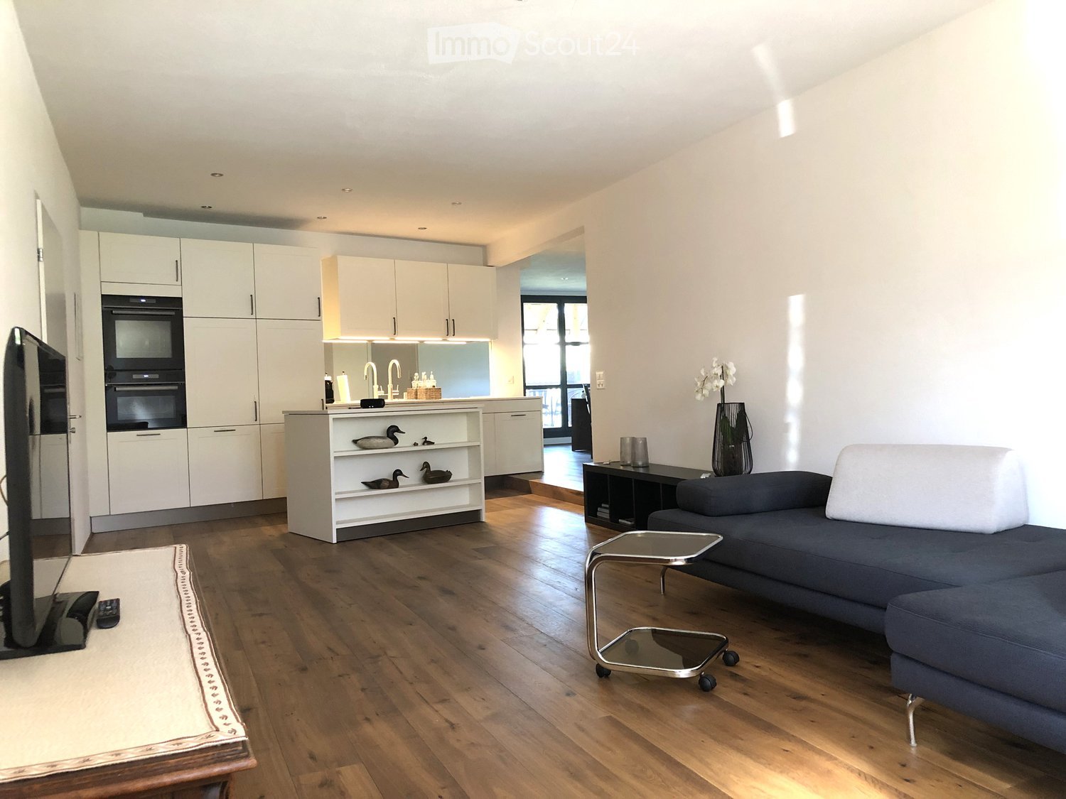 Open-plan living and dining area with a grey sofa, coffee table, television, and a kitchen area with white cabinets and an oven. The space features wooden flooring and large windows.