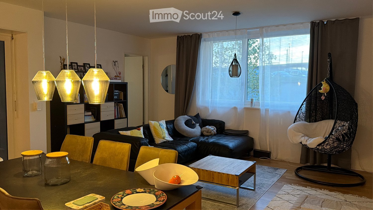 A modern living and dining area with a black leather sofa, colorful cushions, a glass dining table with yellow chairs, a wicker hanging chair, and pendant lights.