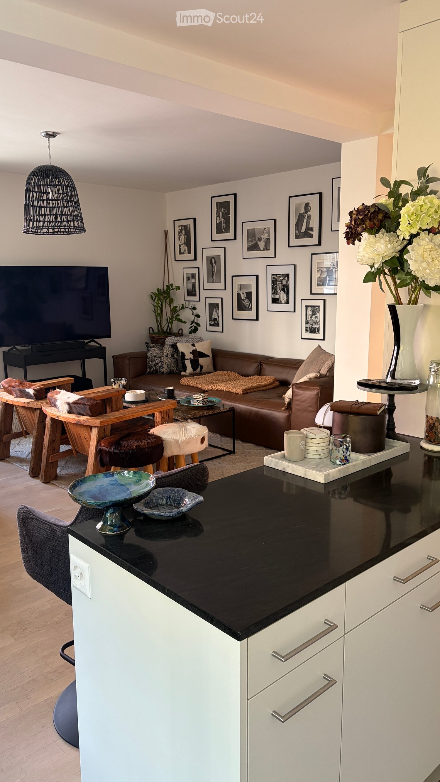 Modern living room with a brown leather sofa, wooden coffee table, decorative bowls, and a pendant light. The space includes a kitchen area with white cabinets and a black countertop.