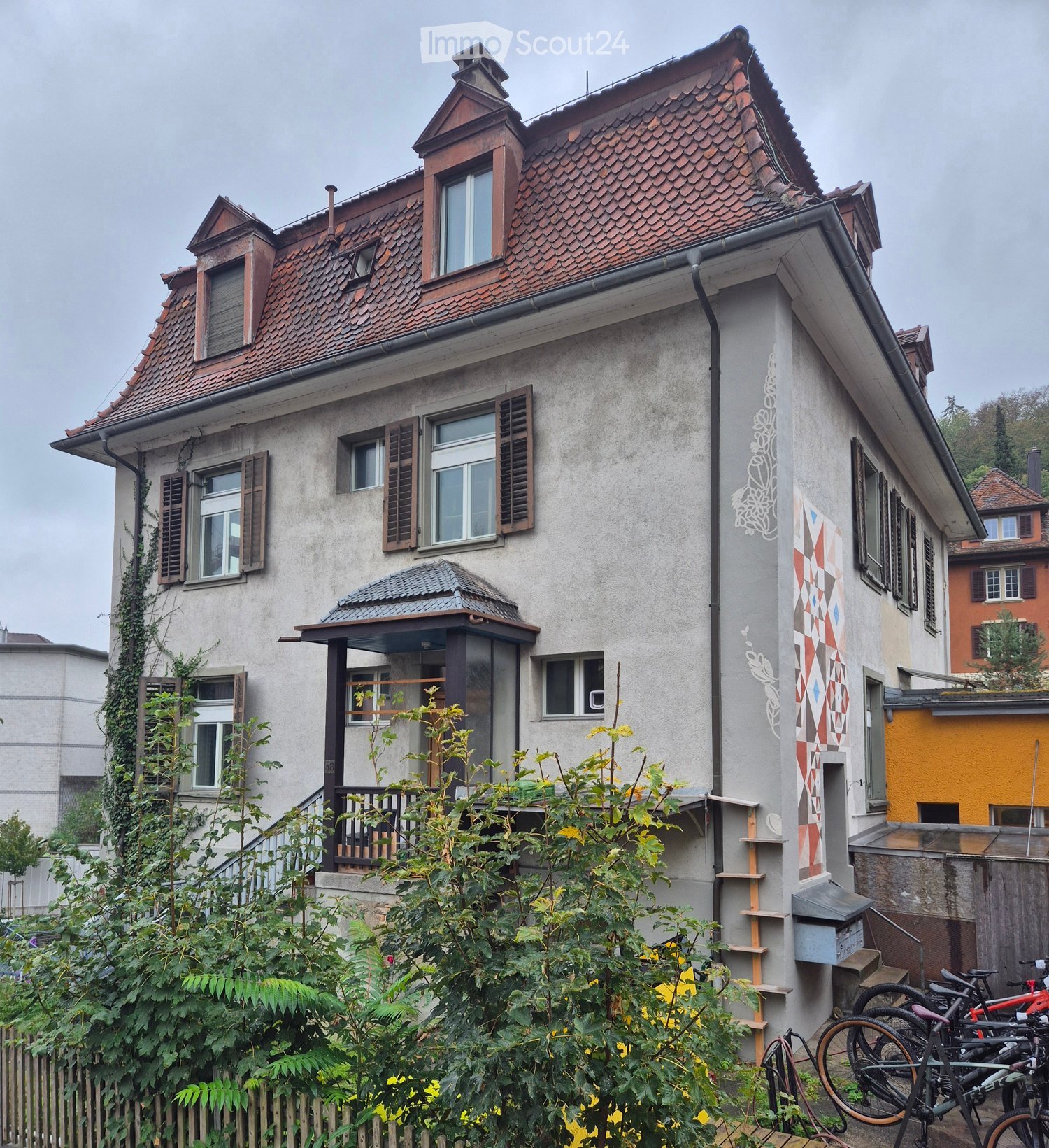 Two-story house with a red-tiled roof, multiple windows with shutters, and a small balcony. The facade features decorative wall art and a small garden with bicycles parked nearby.