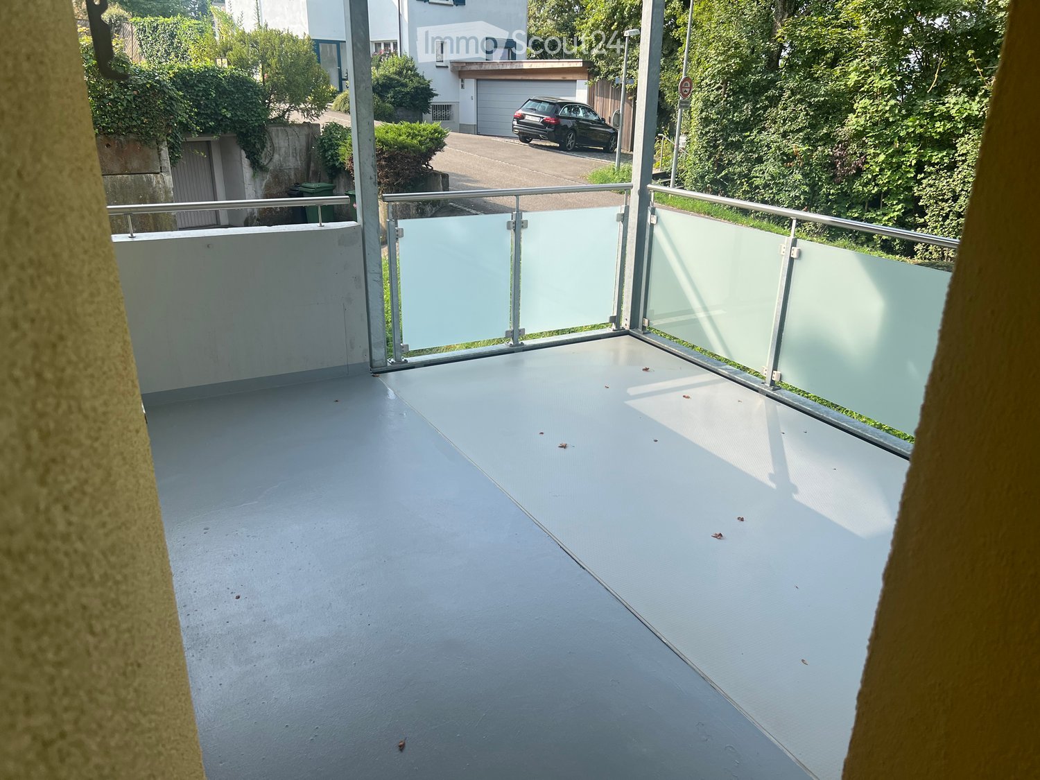 Modern balcony with a smooth grey floor and a glass railing, overlooking a residential area with a parked car and greenery.