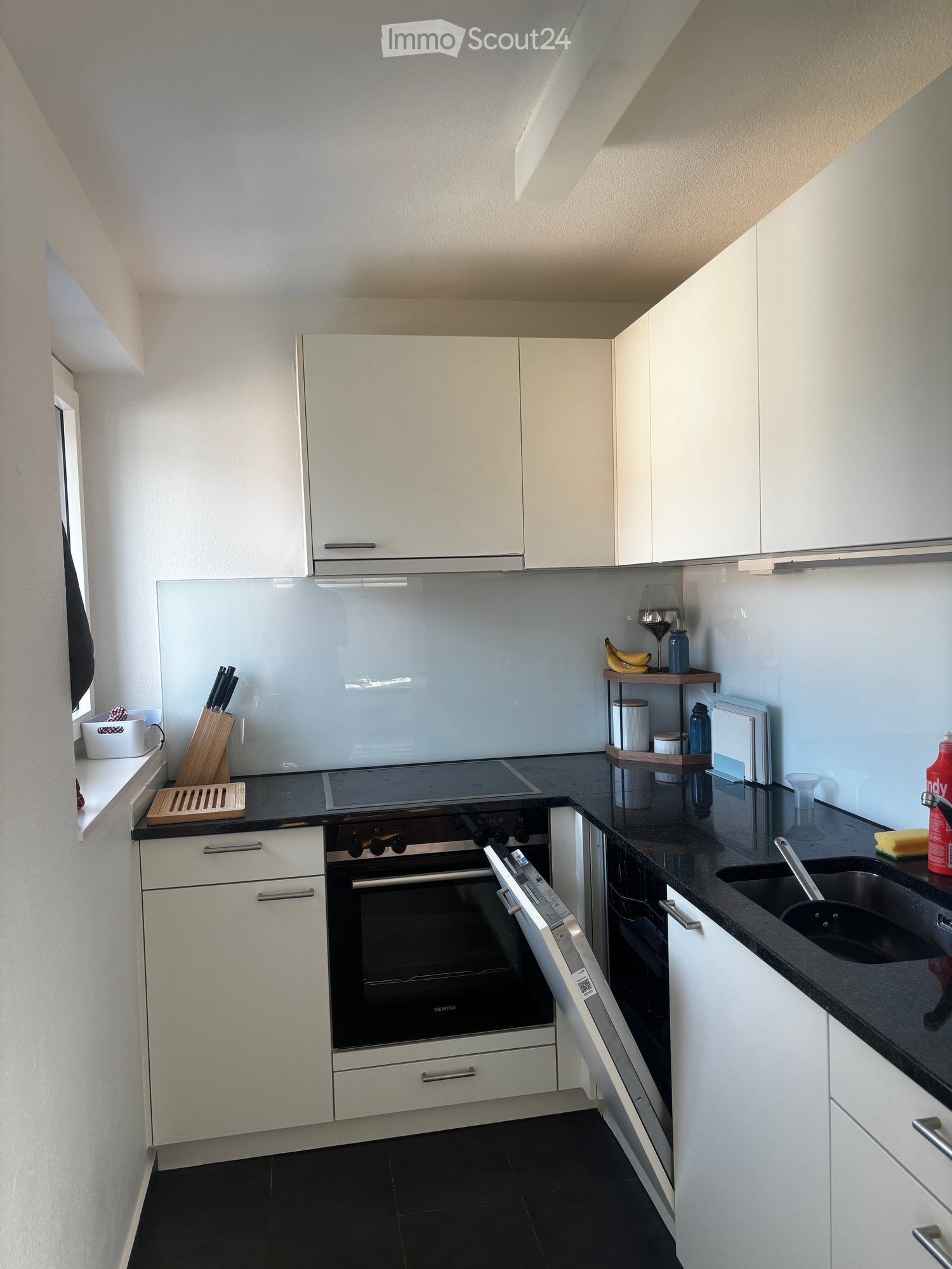 Modern kitchen with white cabinets, black countertops, and stainless steel appliances including an oven, sink, and faucet. A knife block, cup, wine glass, bottle, and frying pan are visible on the countertops.