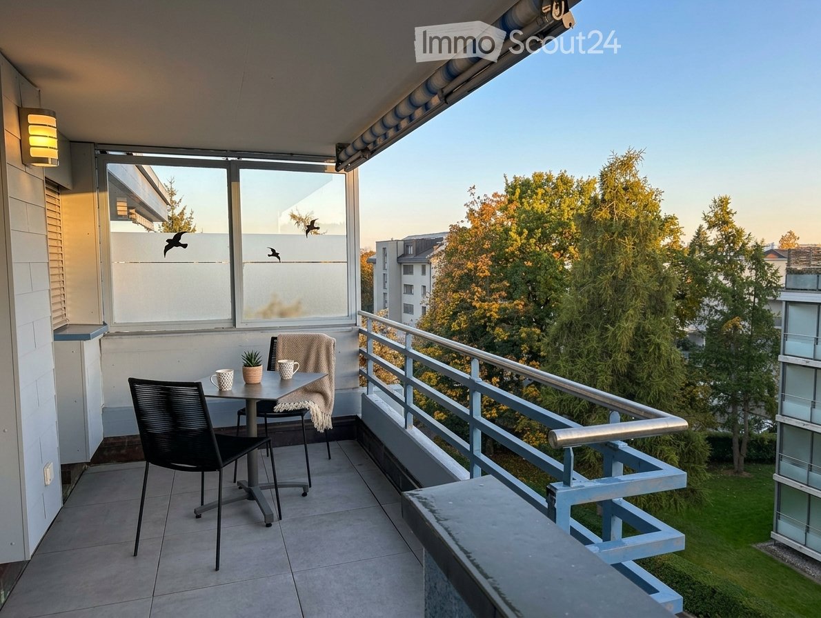 Balcony with a small table, two chairs, a cup, and a small plant, overlooking a green area with trees.
