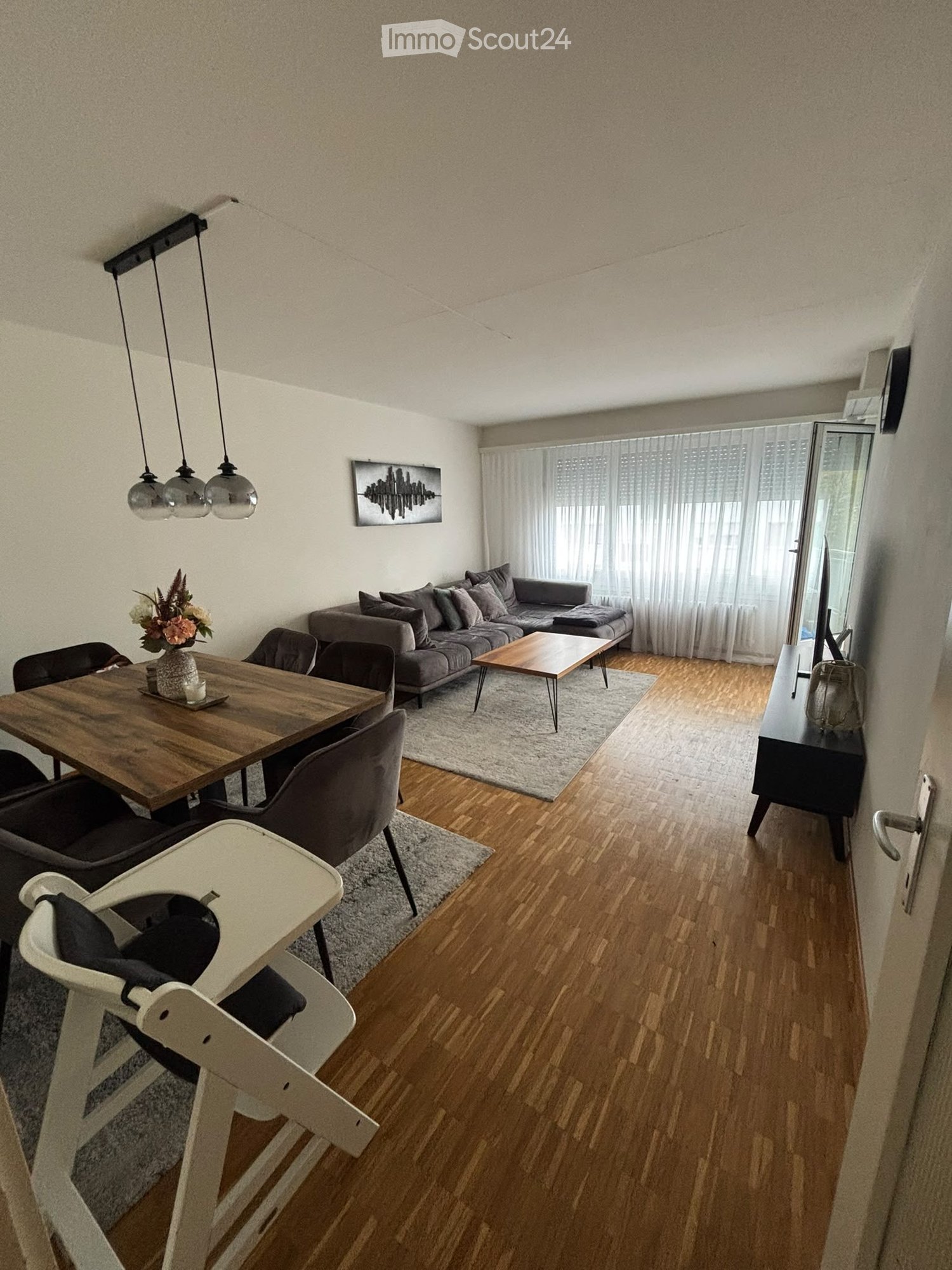 Modern living and dining area with a grey sectional sofa, wooden coffee table, dining table with chairs, baby chair, and decorative elements including a flower vase and artwork.