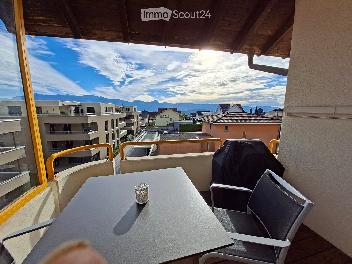 Balcony with a table, chairs, and a cup, overlooking a scenic view of mountains and buildings.