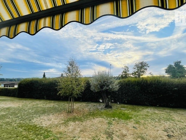 A well-maintained garden with trimmed hedges, olive trees, and a striped awning providing shade. The lawn is green, and the sky is partly cloudy.