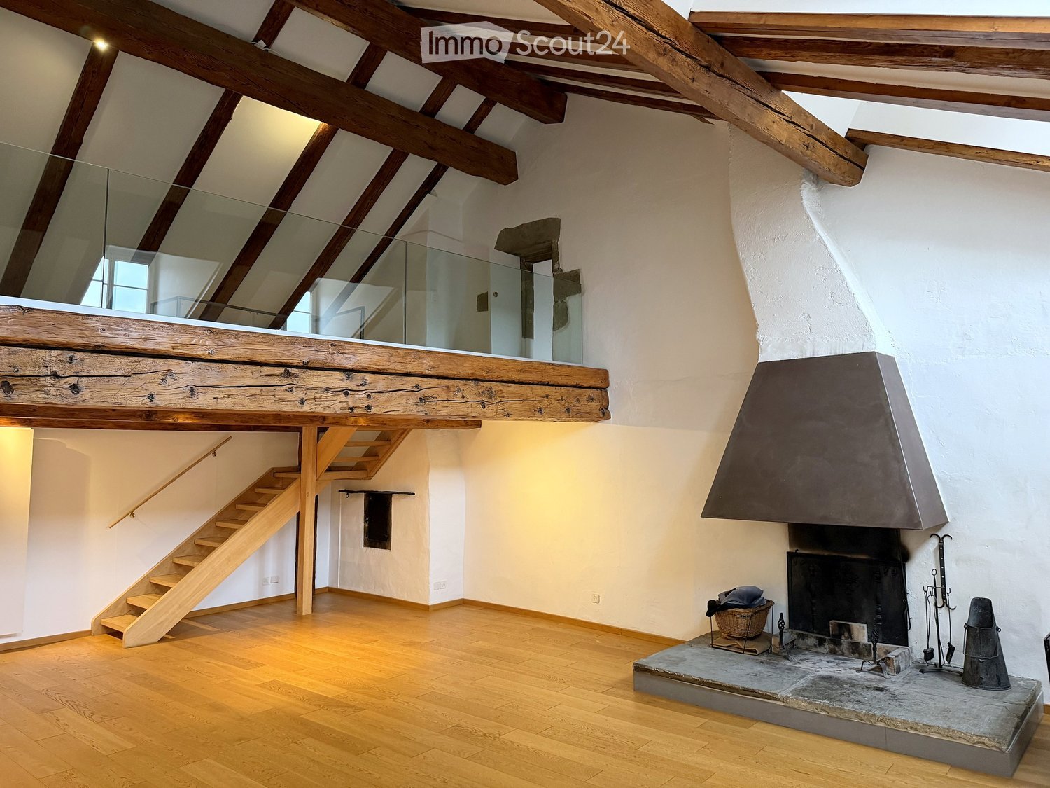 Open living space with wooden beams, exposed wooden staircase, and a stone fireplace with tools and a basket nearby.