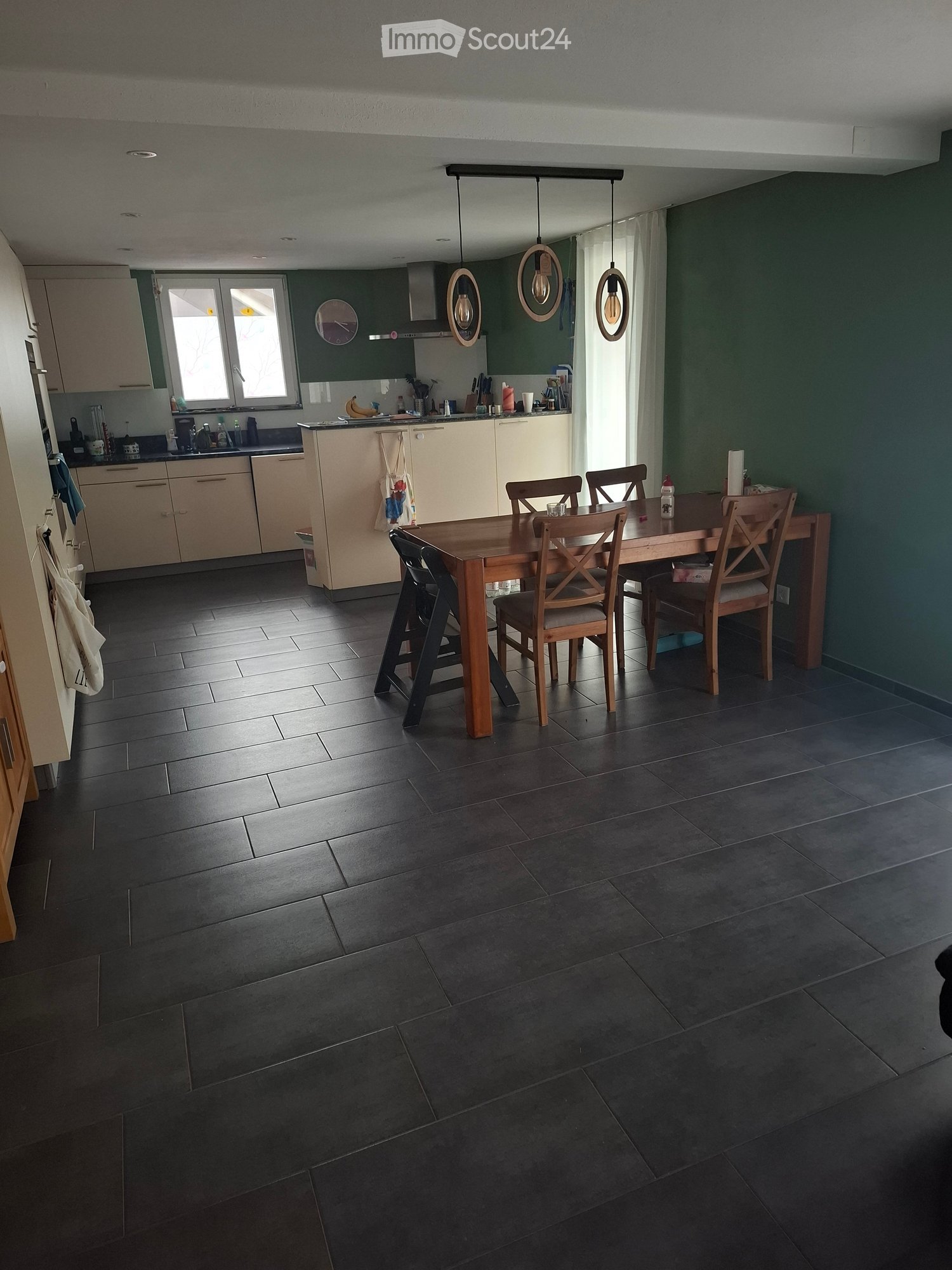 Modern kitchen with dark tiled flooring, light-colored cabinets, and a central dining table with chairs. The kitchen features a sink, refrigerator, and various appliances.