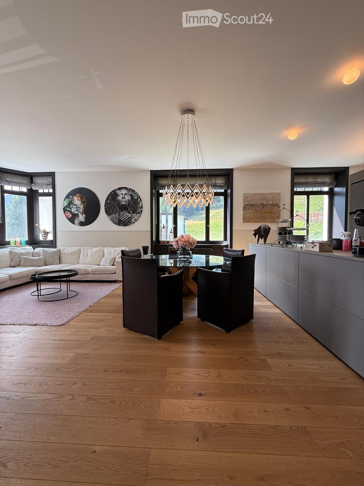 Open-plan living and dining area with light wooden flooring, white sofa, glass dining table, modern chandelier, and large windows.