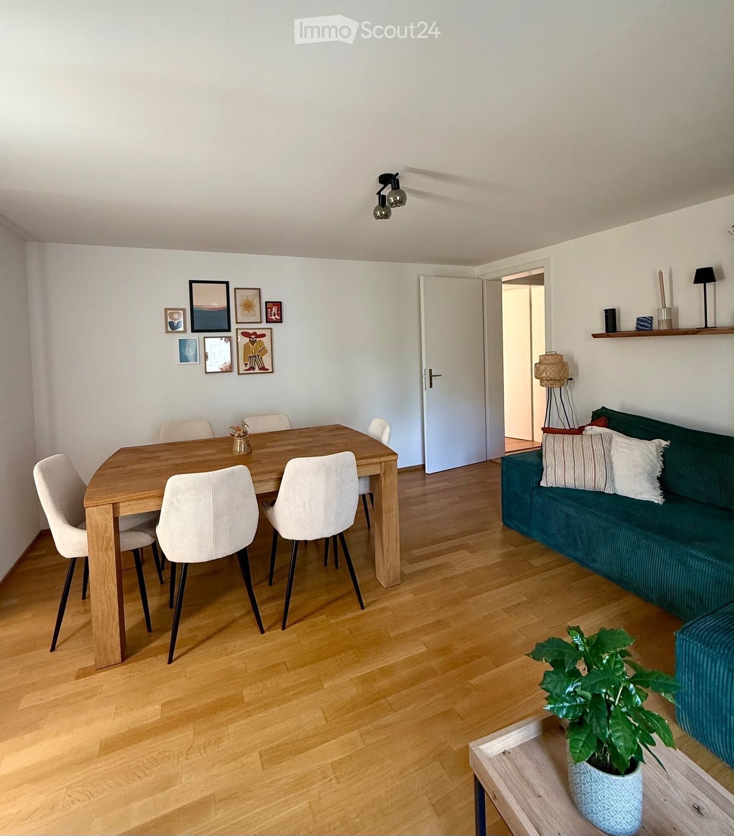Modern living and dining area with a green sofa, wooden dining table, white chairs, and a potted plant on a side table.