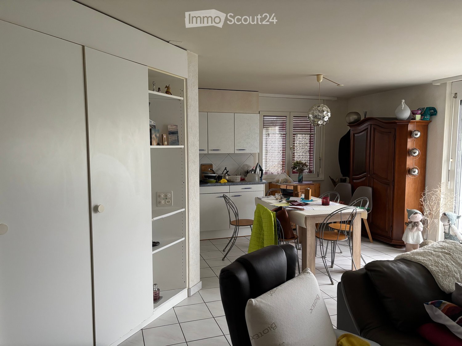 A modern, open-concept living and dining area with a kitchen in the background. The living area features a black leather sofa with white and red pillows, a wooden coffee table, and a dining table with chairs. The kitchen has white cabinets, a countertop w