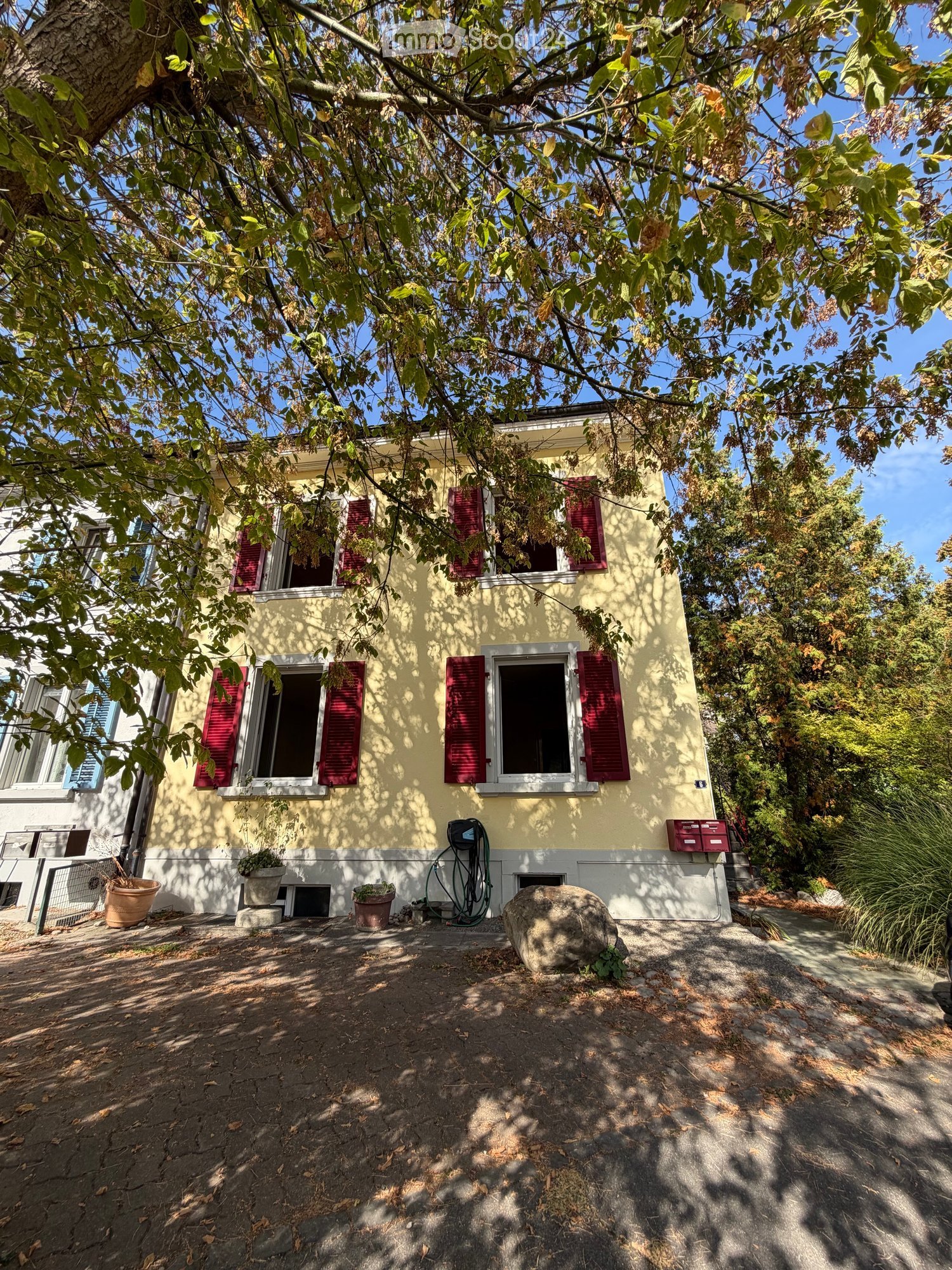 Two-story yellow house with red shutters, surrounded by trees and greenery, with a small garden area and potted plants near the entrance.