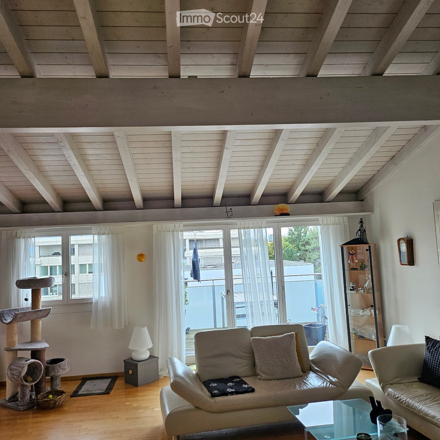 Bright living room with wooden floors, exposed wooden beams, large windows with sheer curtains, and a balcony. Features a beige sofa, armchair, glass coffee table, cat tree, and a small lamp.