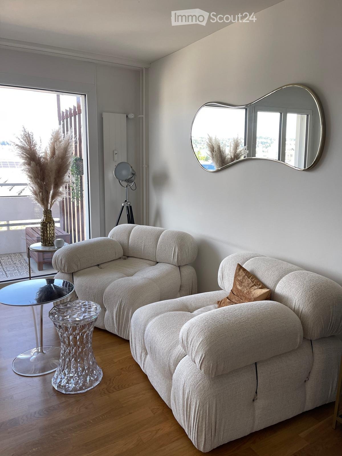 Modern living room with a light grey sectional sofa, glass coffee table, crystal stool, decorative vase with dried plants, and a large round mirror on the wall.
