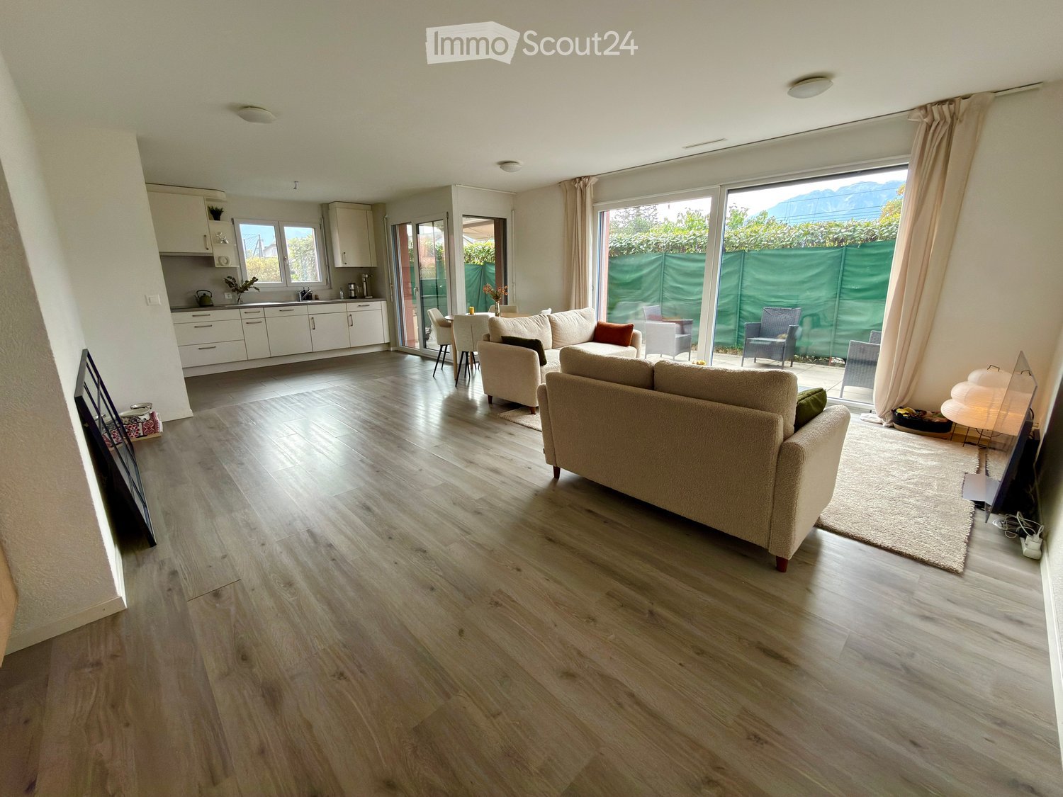 Open-plan living and dining area with light wooden flooring, cream-colored sofa, dining table with chairs, and a modern kitchen with white cabinets.