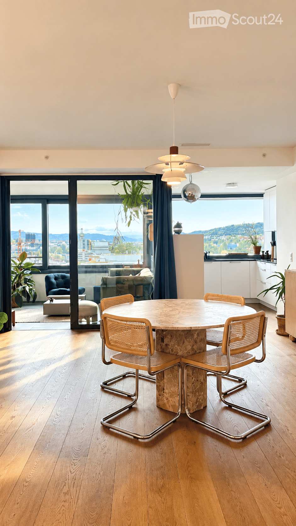 Modern dining area with a round marble table, four wicker chairs, pendant lighting, and a view of the cityscape through large windows. A small seating area with a sofa and ottoman is visible through the open doorway.