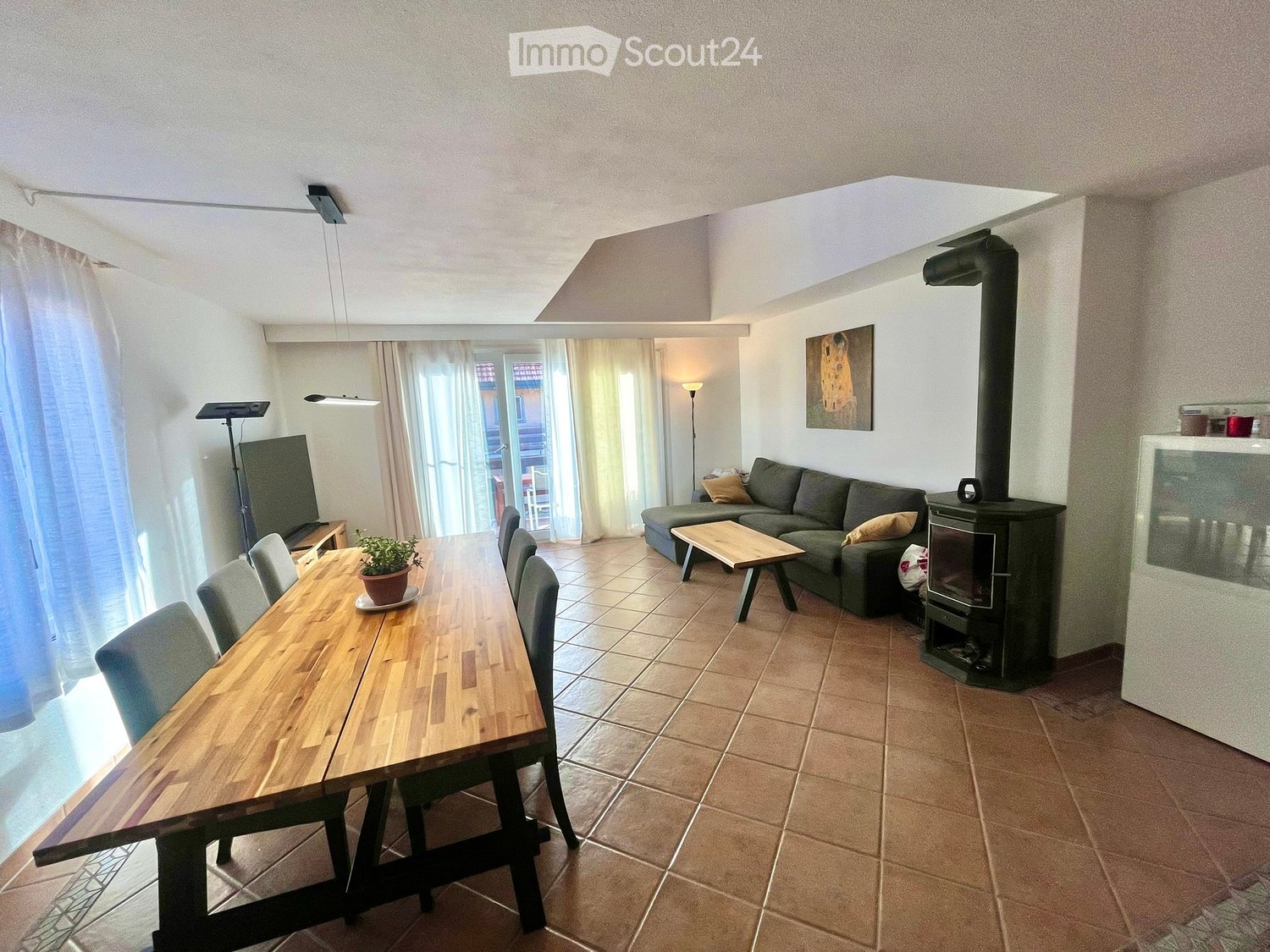 A modern living and dining area with a wooden dining table, chairs, a sofa, a small wooden stool, a fireplace, and a television. The space is well-lit with large windows and a skylight.