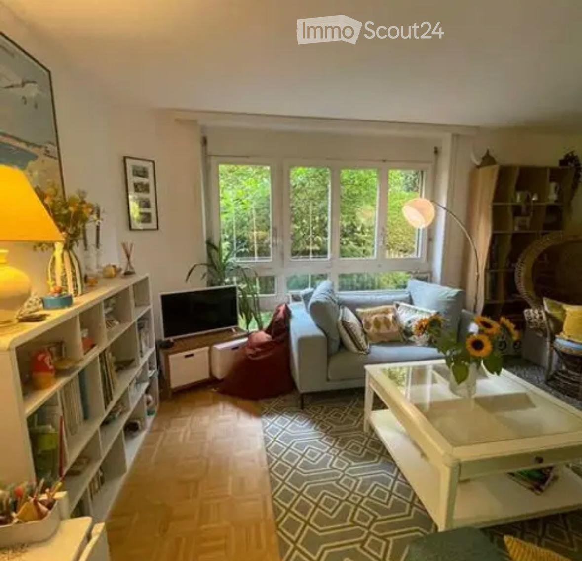 Bright living room with large windows, light grey sofa, coffee table, bookshelf, floor lamp, and decorative items.