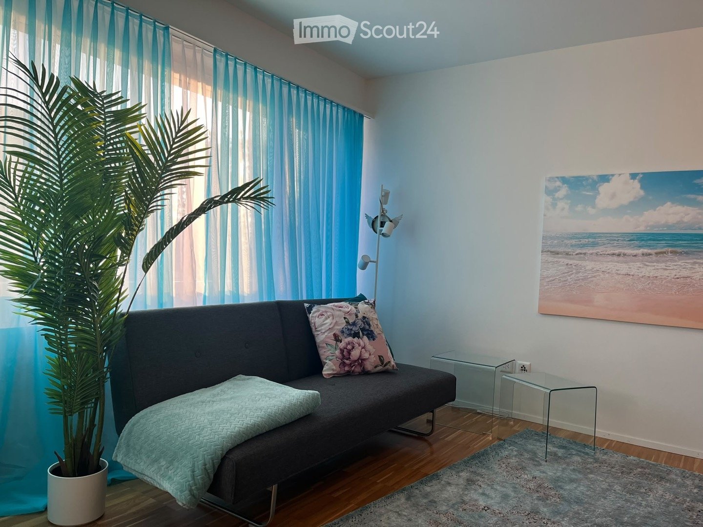A modern living room with a grey sofa, decorative pillows, a small glass coffee table, a floor lamp, a large painting of a beach scene, and a potted plant near the window with blue curtains.