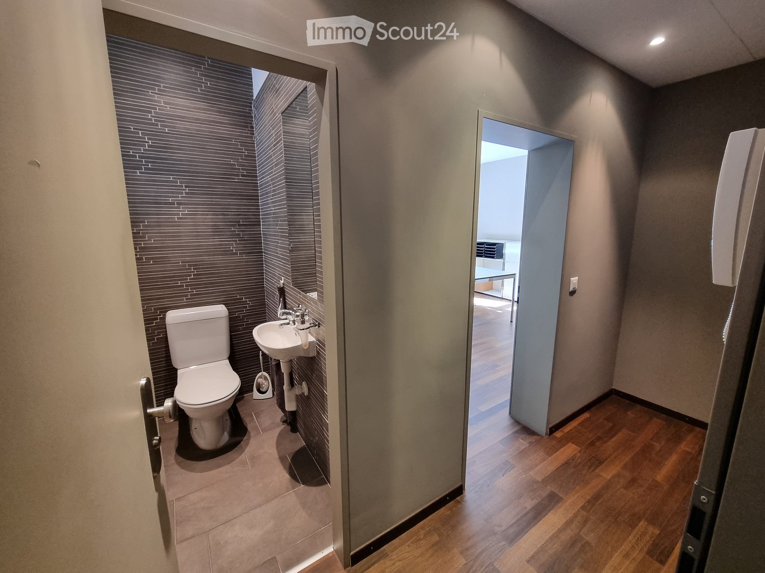 Modern bathroom with a toilet, sink, and faucet, featuring dark tiled walls and a wooden floor visible through an open doorway.