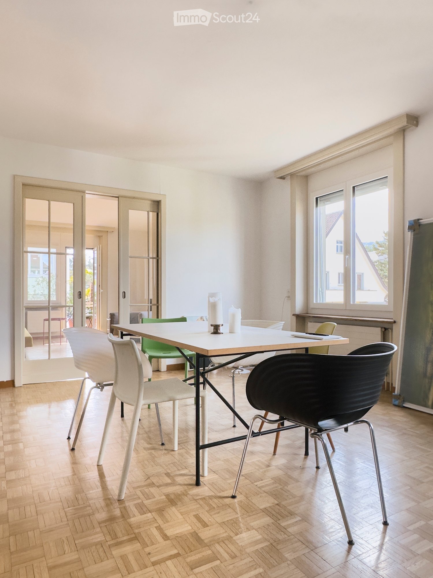 Bright room with wooden herringbone flooring, white table with four chairs, and a candle centerpiece. Large windows and sliding glass doors lead to a balcony area.