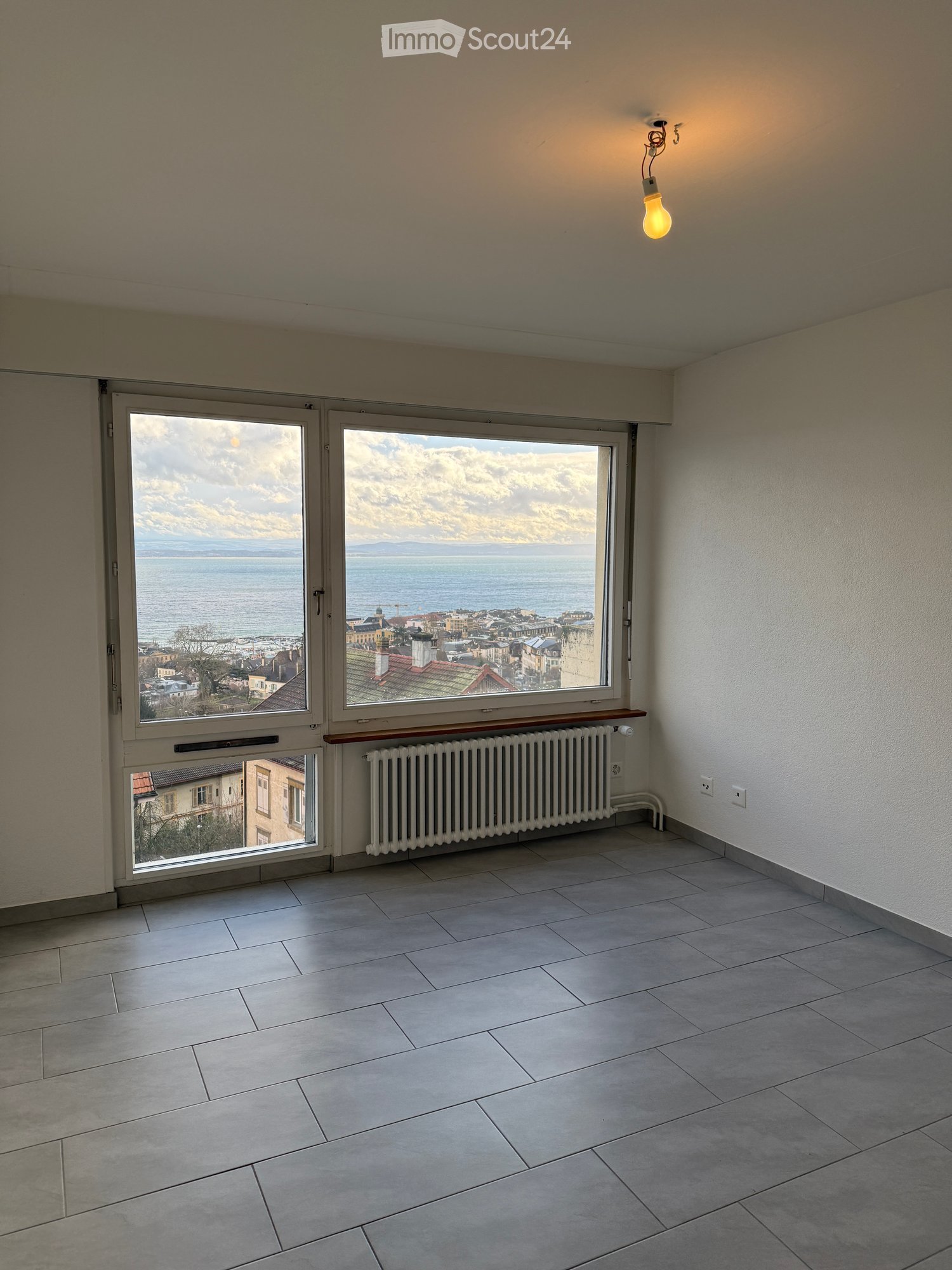 Empty room with tiled floor, large windows offering a scenic view of a city and body of water, radiator below the windows, and a single light fixture on the ceiling.