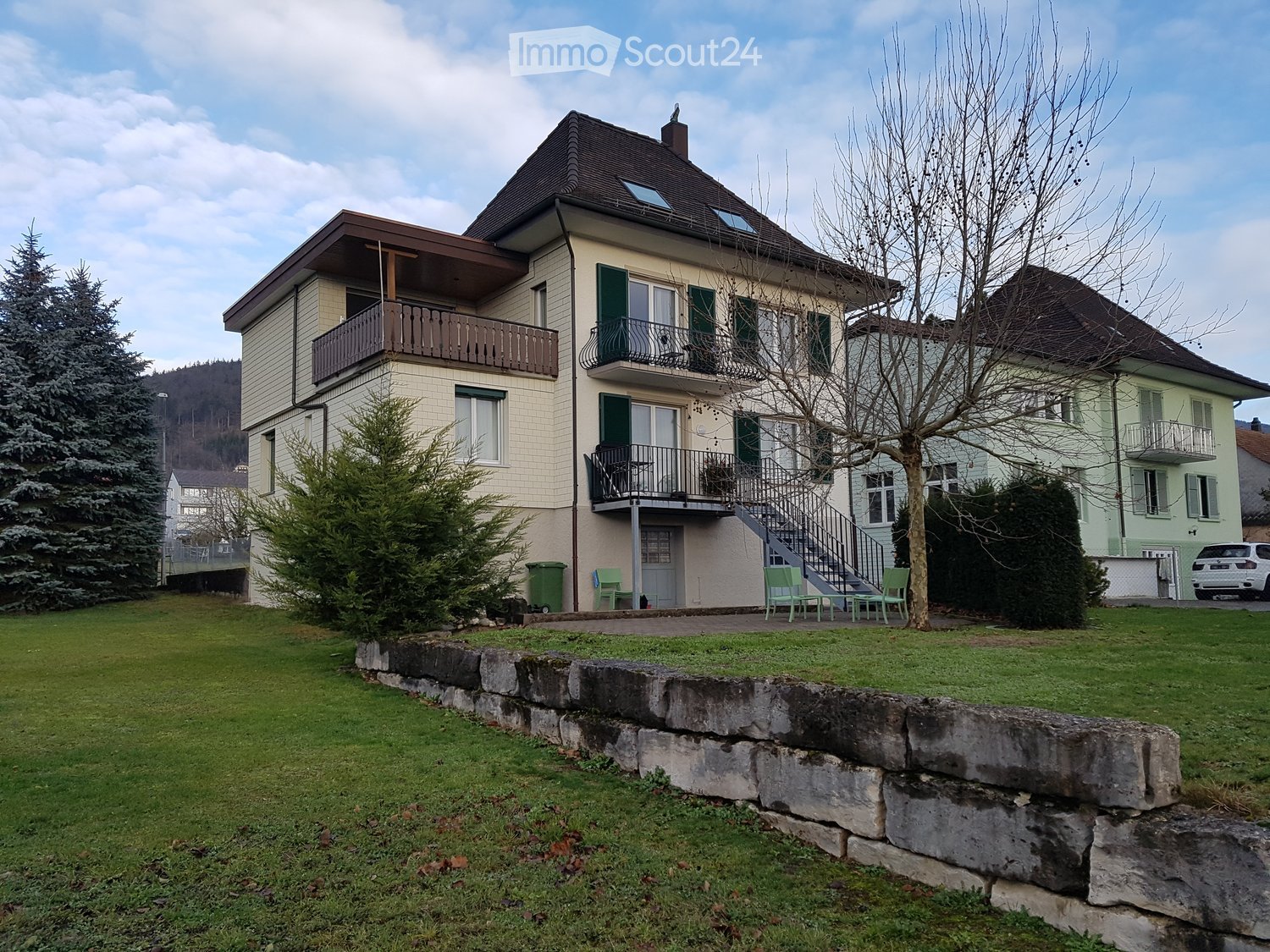 Two-story house with beige siding, green shutters, multiple balconies, and a well-maintained lawn.