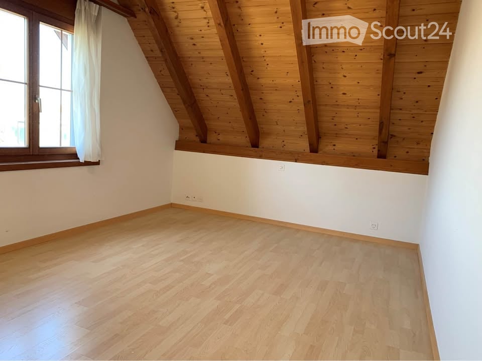Empty room with wooden flooring, white walls, and exposed wooden beams on the ceiling. A window with white curtains is visible on one side.