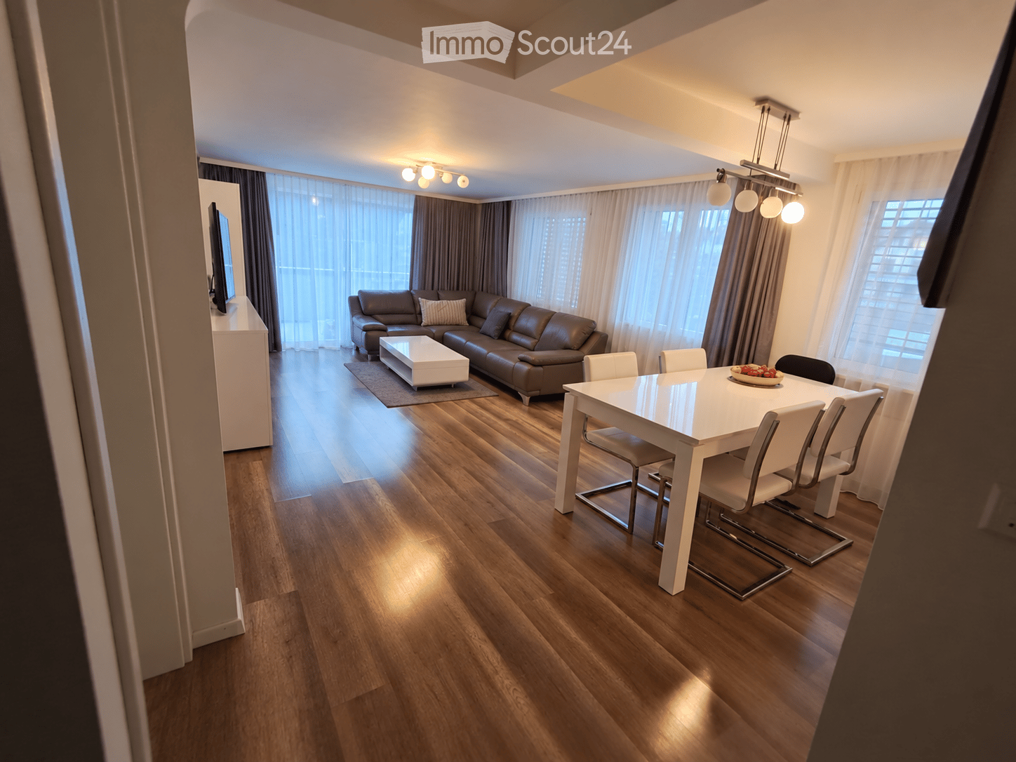 Modern living and dining area with wooden flooring, large windows with curtains, a brown leather sofa, a white coffee table, a white dining table with six chairs, and a TV on a white stand.