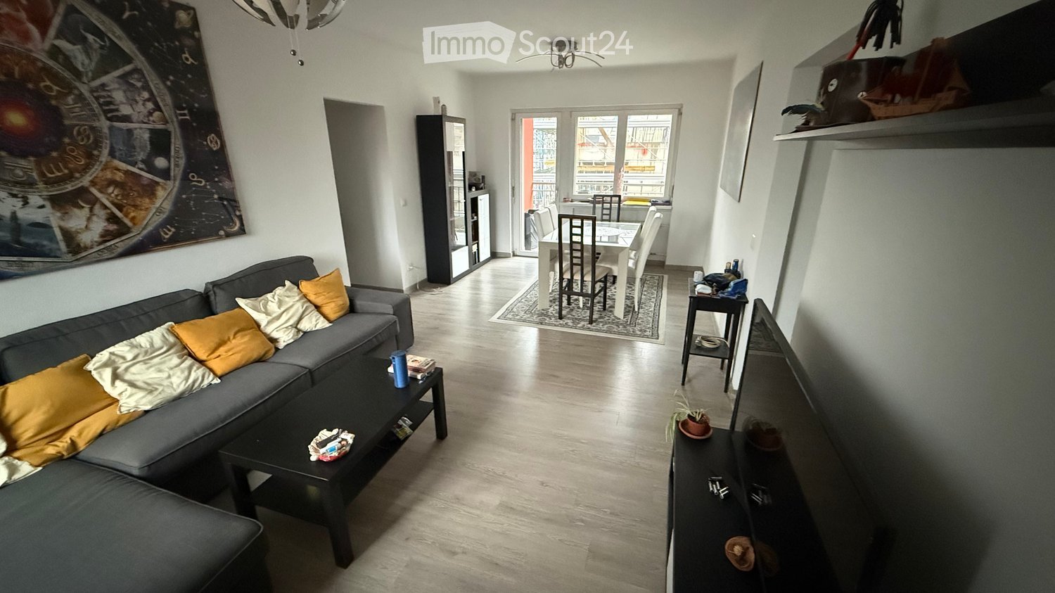 Open-plan living and dining area with a grey sectional sofa, coffee table, dining table with chairs, and a large window leading to a balcony.