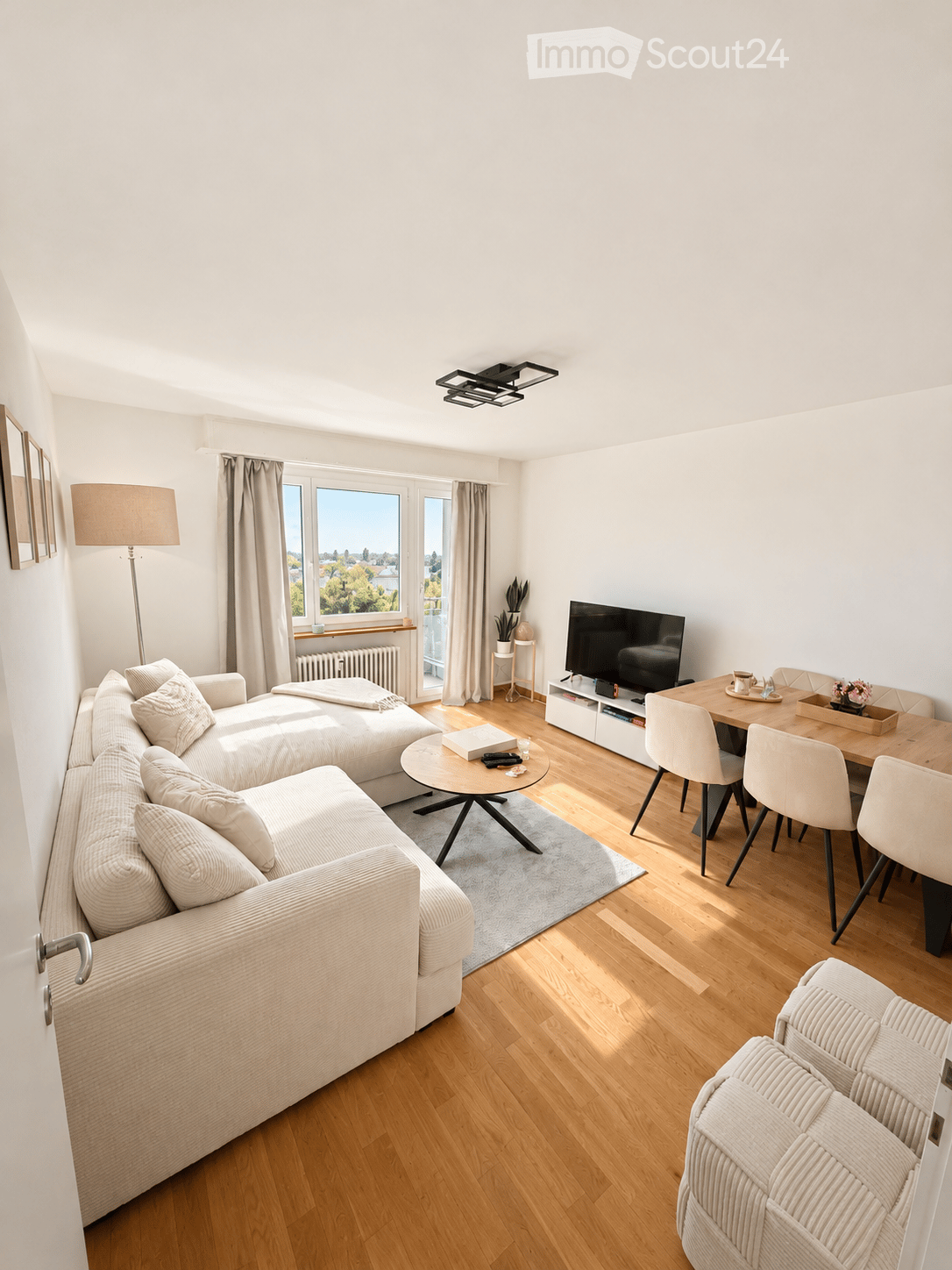 Modern living room with a large window offering a view, featuring a sectional sofa, coffee table, dining table with chairs, TV, floor lamp, and a small plant on a side table.