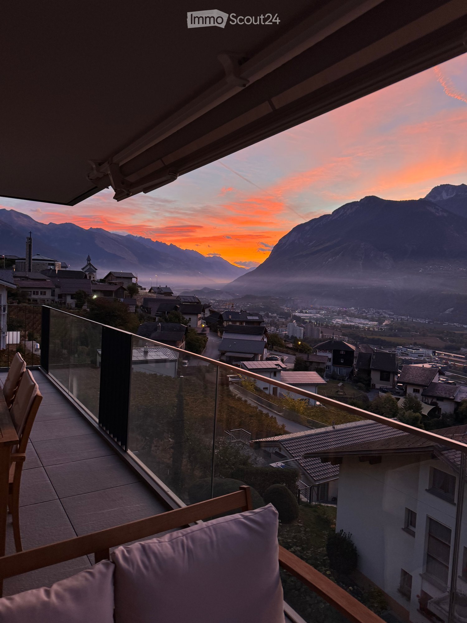 Balcony with glass railing, wooden chairs, and a table, overlooking a town and mountains at sunset.