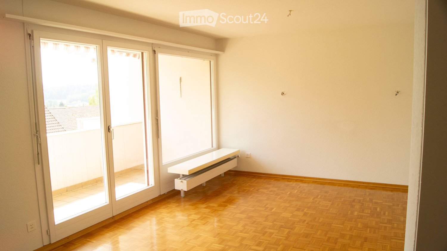 Empty room with wooden flooring, large sliding glass doors leading to a balcony, and a built-in radiator bench.