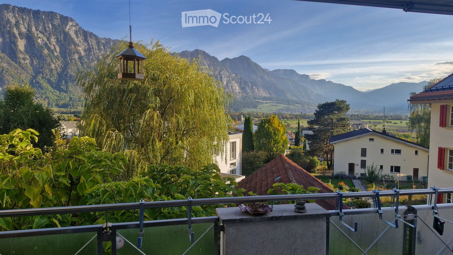 Balcony overlooking a scenic valley with mountains, trees, and houses in the background.