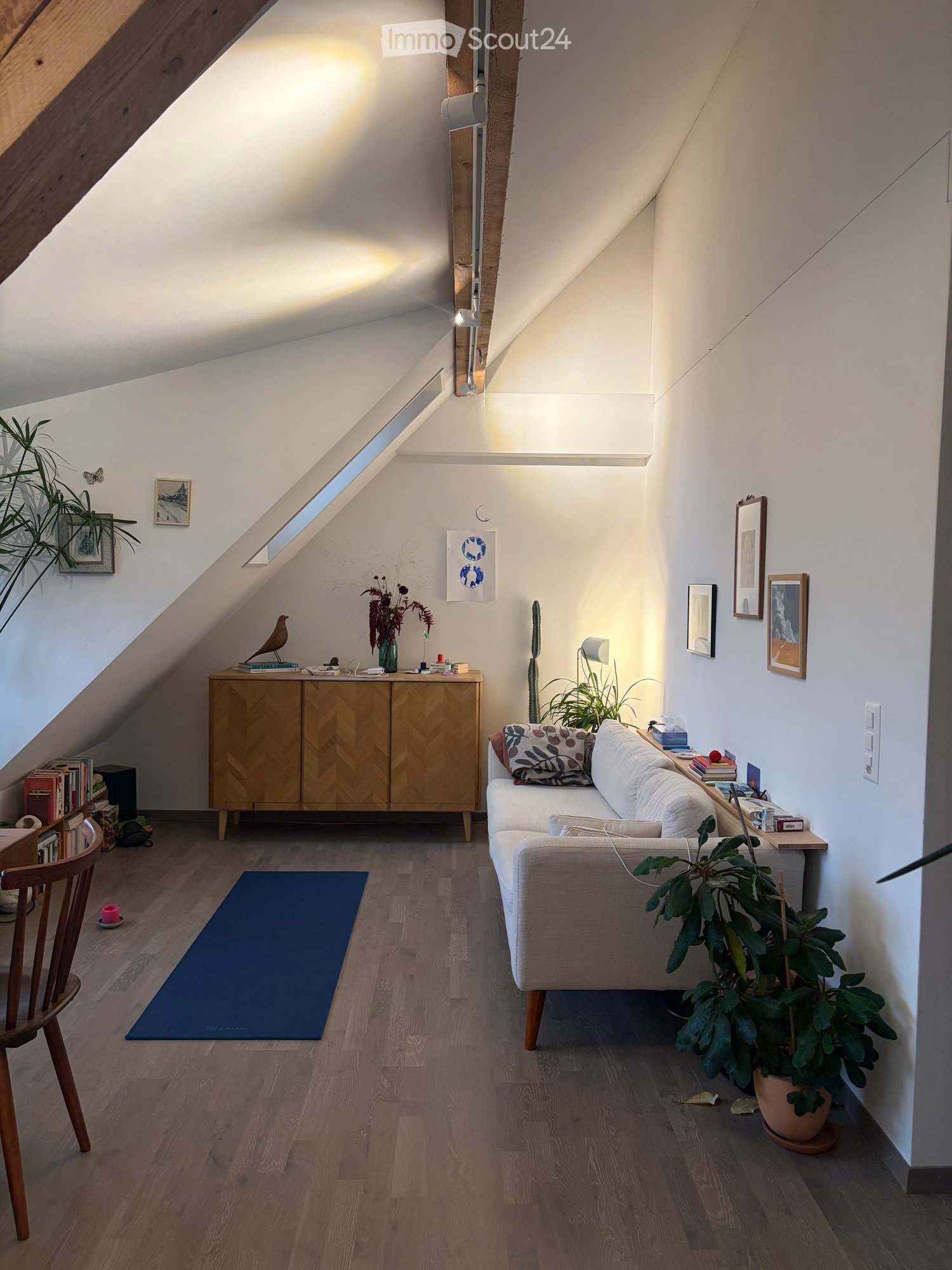 Cozy attic living room with wooden floors, slanted ceilings, and large windows. Features a sofa, wooden cabinet, chair, small table, and several plants.