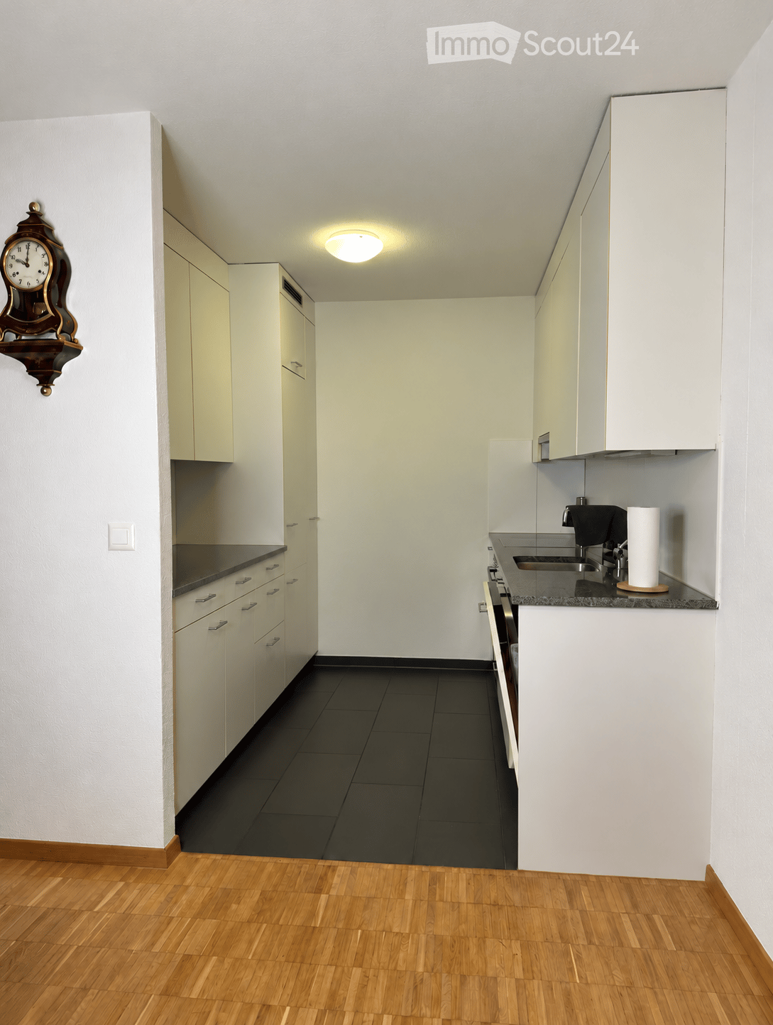 Modern kitchen with white cabinets, black countertops, and a dark tiled backsplash. Features a sink, faucet, oven, and a wall-mounted clock.