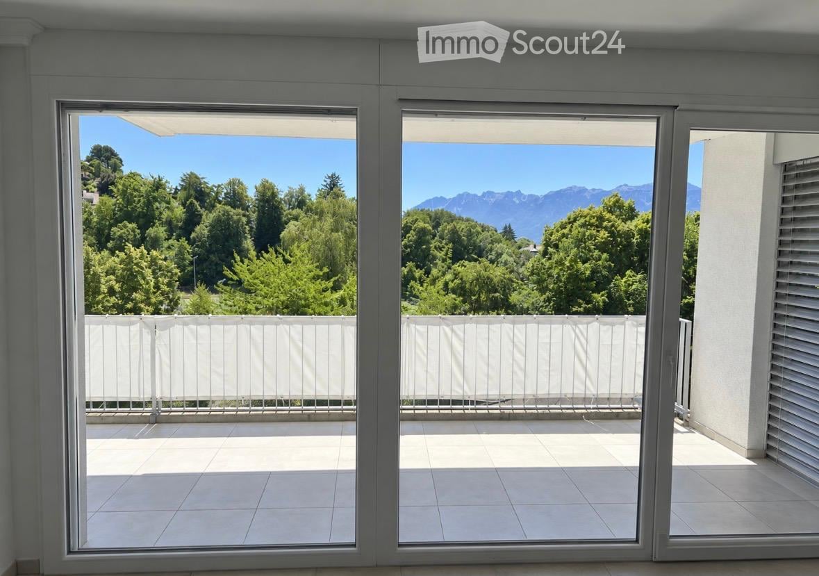 Large, modern balcony with white tiled flooring, white metal railing, and expansive views of green trees and distant mountains.
